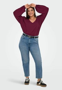 Weinrotes Langarmshirt mit V-Ausschnitt, kombiniert mit hoch taillierten blauen Jeans. Schwarzer Gürtel mit silberner Schnalle, dazu schwarze und weiße Sneakers.