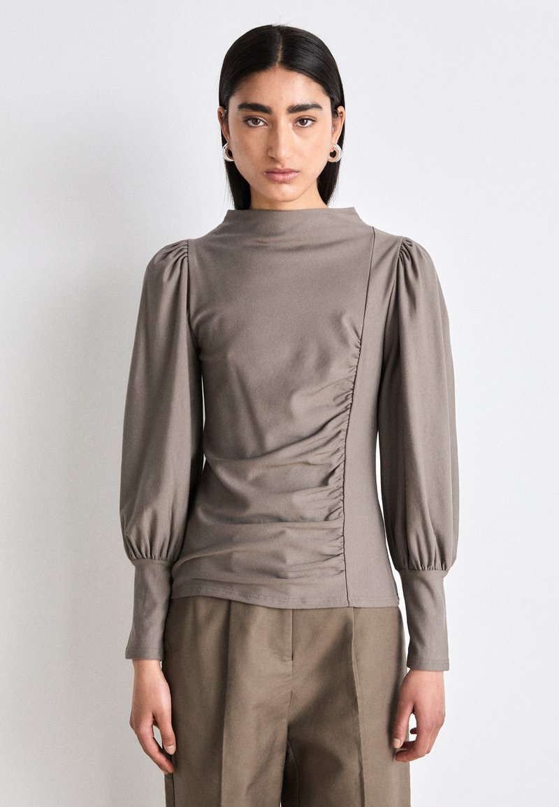 Haut à manches longues et col montant en tissu taupe avec détail froncé sur les côtés et manches volumineuses. Assorti à un pantalon large taille haute.