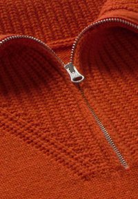 Pomarańczowy sweter z dzianiny z pół zamkiem błyskawicznym, z wystającymi wzorami teksturowymi oraz gładkimi sekcjami. Metalowy zamek błyskawiczny wyraźnie wyeksponowany.