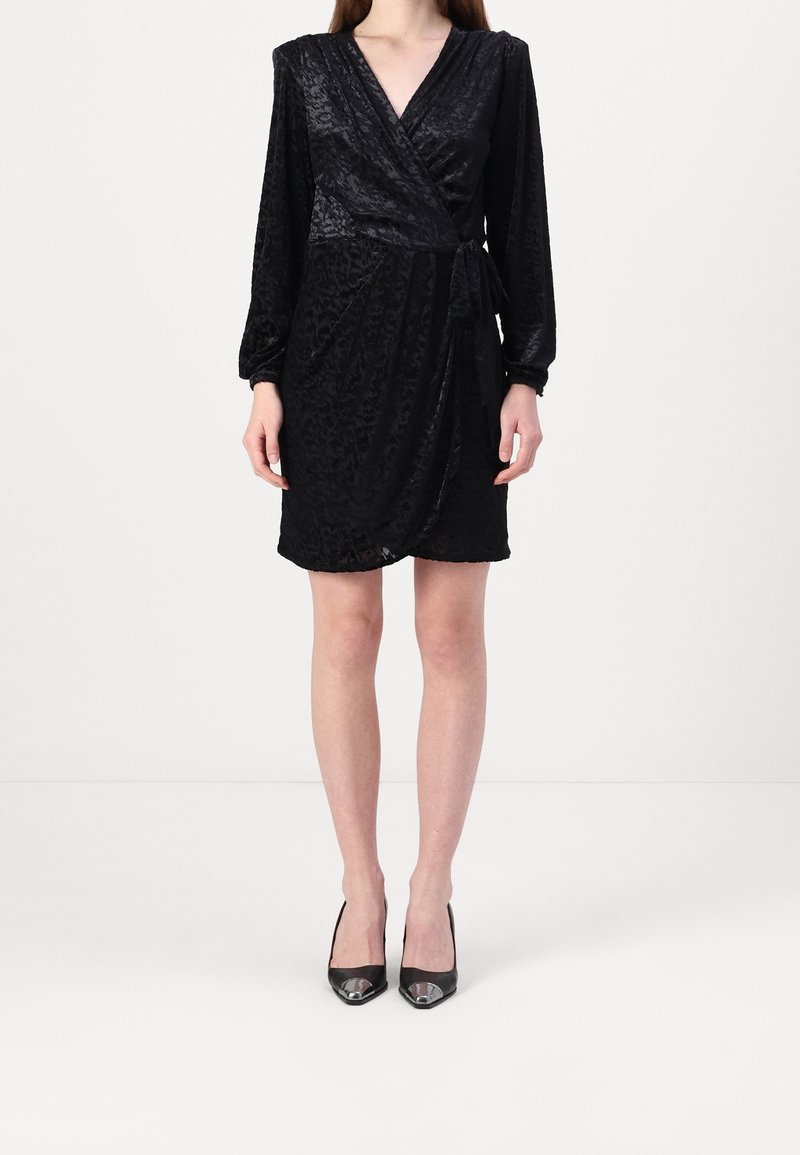 Femme portant une robe portefeuille noire texturée à manches longues et des talons hauts noirs à bouts pointus, debout devant un fond blanc uni.