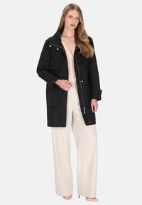 Sort funktionel jakke med lynlåslukning, flere lommer foran og høj krave. Kombineret med cremefarvede bukser med vidde i benene.