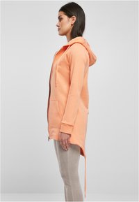 Hoodie v broskvofarebnej farbe s predným zipsom, klokanymi vreckami a dlhším zadným lemom, s dlhými rukávmi a textúrovanou tkaninou.