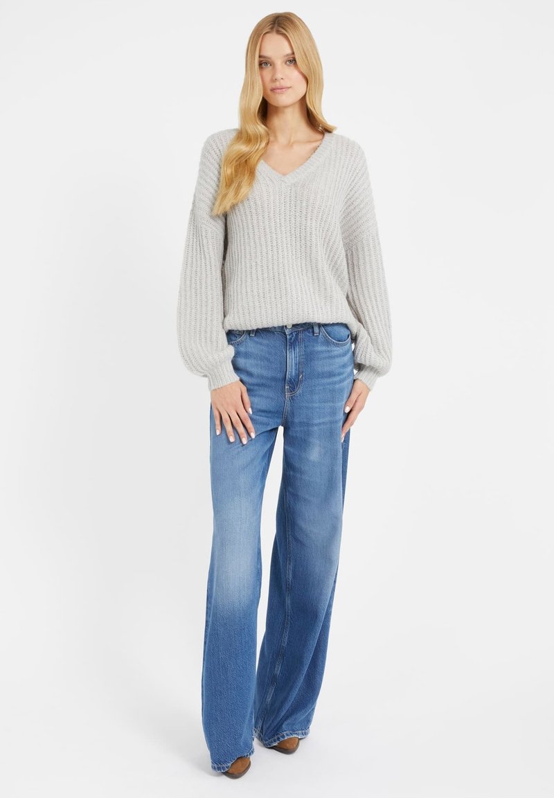 Heller grauer Strickpullover mit V-Ausschnitt und gerippter Textur, kombiniert mit hellblauen weit geschnittenen Jeans und braunen Stiefeletten mit Absatz.