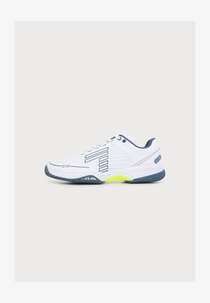 Baskets de sport blanches avec semelle bleu marine, bordure noire et accent jaune, vue de profil avec détails de la marque sur fond uni.