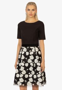 Schwarzes Kleid mit enganliegendem Oberteil und kurzen Ärmeln, das einen weißen, floralen Spitzenrock und ein semi-transparentes Überkleid zeigt, verziert mit einem Taillenbund.