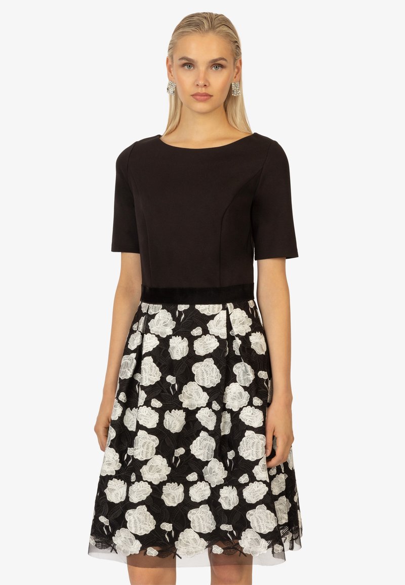 Schwarzes Kleid mit enganliegendem Oberteil und kurzen Ärmeln, das einen weißen, floralen Spitzenrock und ein semi-transparentes Überkleid zeigt, verziert mit einem Taillenbund.