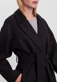 Cappotto in misto lana nero con ampi risvolti, vita cinturata e tasche laterali; presenta una morbida texture e una vestibilità comoda.