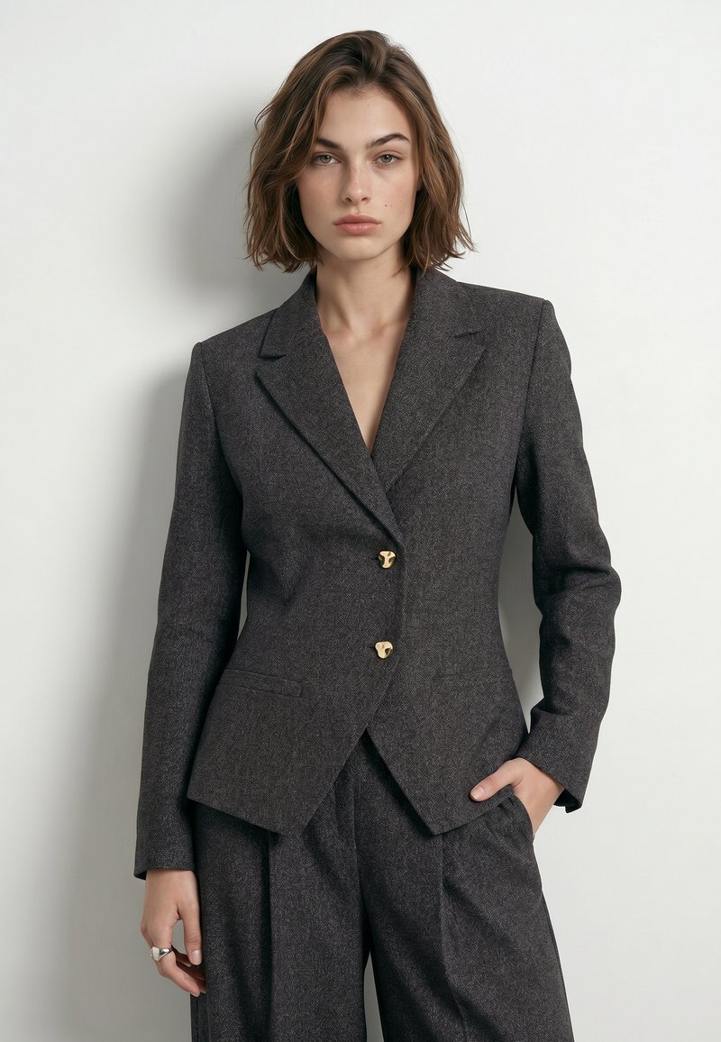 Femme aux cheveux courts et bruns portant un blazer gris foncé cintré avec des boutons dorés et un pantalon assorti, debout devant un fond clair uni.