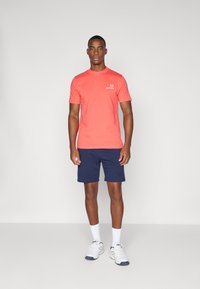 Korallfarget T-skjorte med kort erm og logo, sammen med marineblå shorts. Modellen har på seg hvite joggesko med grå detaljer og hvite sokker.