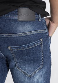 Vaqueros de denim en un lavado azul oscuro con una textura desvaída. Las características incluyen un parche de cuero negro con "GIANNI KAVANAGH" grabado y dos bolsillos traseros.