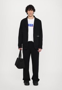 Veste en laine noire avec revers, t-shirt blanc avec graphique bleu, pantalon noir, sac noir et chaussures noires massives.