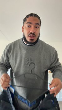 Pelēks sporta krekls ar reljefa logotipu, turēts uz melnas pakaramās. Melnas bikses daļēji redzamas, ar zilu jostas joslas etiķeti.