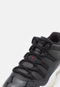 Černá kožená teniska s texturovaným povrchem, vybavená černými tkaničkami a červeným logem. Zaoblená špička a polstrovaný límec kolem kotníku.