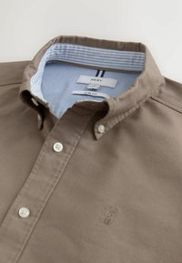 Brun slim fit skjorte med knaplukning, hvide knapper, stribet indvendig krave og lille broderet hjortelogo på brystet.