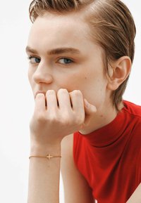 Pulsera de oro con un charm de cruz, llevada en una mano que reposa bajo la barbilla. La persona tiene el cabello corto y viste una blusa roja de cuello alto.