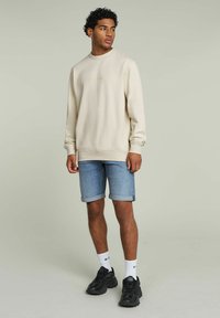 Sudadera beige con cuello redondo y bordado, combinada con pantalones cortos de mezclilla azul claro doblados en el dobladillo, zapatillas negras y calcetines blancos.