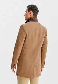 Brązowy płaszcz z wełny w składzie, o klasycznym kroju, z ciemnym kołnierzem, guzikami na rękawach oraz gładkim, ustrukturyzowanym wykończeniem.