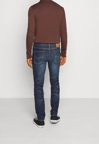 Hombre con jeans azul oscuro, camisa de manga larga marrón y zapatillas Converse de lona negras de corte bajo, posando frente a un fondo liso.