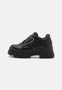 Fekete szintetikus bőrből készült sneaker vastag fekete gumi talppal, csattal ellátott részlettel és fűzős szemlyukasztásos kialakítással. Texturált anyag a stabilitásért.