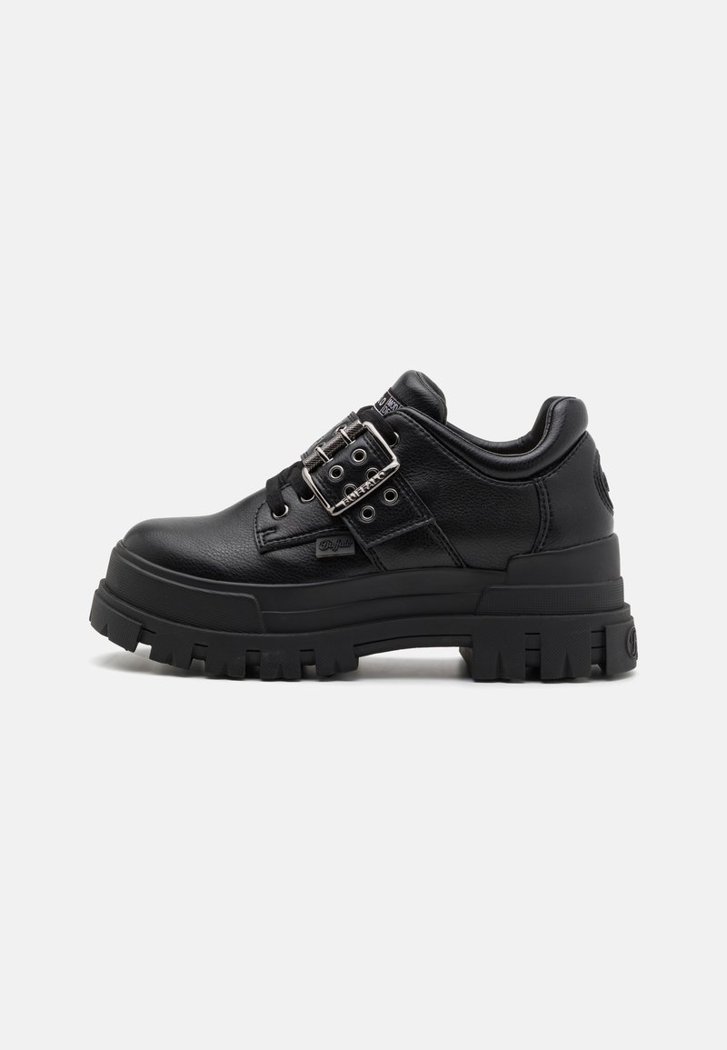 Fekete szintetikus bőrből készült sneaker vastag fekete gumi talppal, csattal ellátott részlettel és fűzős szemlyukasztásos kialakítással. Texturált anyag a stabilitásért.