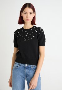 Zwarte gebreide trui met korte mouwen en een geribbelde zoom, versierd met verspreid aangebrachte transparante edelstenen op de schouders. G gedragen met lichtblauwe denim jeans.