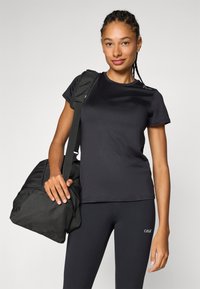 Camiseta atlética negra de manga corta, con textura suave, combinada con leggings negros. Una bolsa de deporte negra es sostenida por el brazo derecho del sujeto.