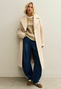 Mujer con un abrigo de lana largo beige, suéter de cuadros, jeans oscuros de pierna ancha y zapatillas marrones, de pie contra una pared clara y lisa.