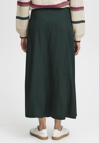 Jupe longue et vert foncé en tissu lisse, avec une fente latérale discrète, assortie à des baskets blanches sur un fond uni.
