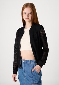 Crna bomber jakna s čipkastim rukavima, prednjim zatvaračem i rebrastim manžetama. Nosí se preko bež kratkog topa, u kombinaciji s traper suknjom koja ima džepove.