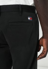 Czarne spodnie o gładkiej fakturze, z tylną kieszenią i naszywką z logo Tommy Jeans w kolorach czerwonym, białym i niebieskim.