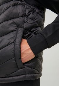 Gilet imbottito nero caratterizzato da una texture trapuntata, tasca laterale e dettagli a costine sui polsini. Il materiale appare liscio e lucido.