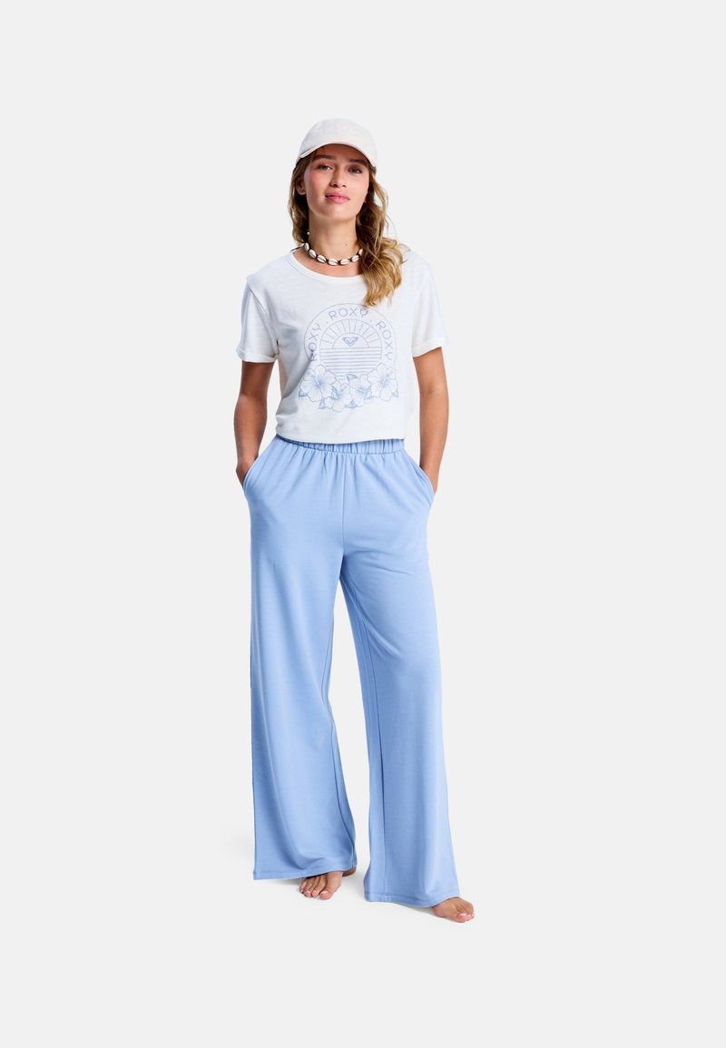 Lichtblauwe wijde broek met een elastische taille, gecombineerd met een witte t-shirt met korte mouwen met een blauwe grafische print. Een witte pet maakt de look compleet.