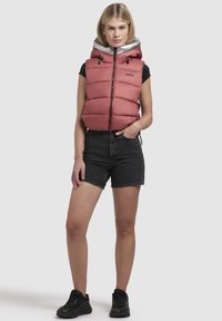 Vest îmblănit în roz pal cu glugă argintie, fermoar frontal și șnururi ajustabile, asortat cu pantaloni scurți din denim negri și adidași negri.