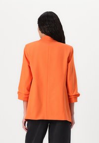 Orange blazer med skræddersyet pasform, trekvart ærmer og en glat tekstur, med et enkelt split bagpå, båret over sorte bukser.