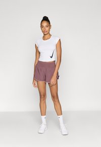 Λευκό κοντό αθλητικό τοπ με μαύρο λογότυπο, σε συνδυασμό με μωβ σορτς με λευκές ρίγες. Το σύνολο ολοκληρώνεται με λευκά αθλητικά παπούτσια.