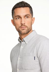 Lichtgrijs overhemd met knoopsluiting en een zak, gemaakt van gestructureerde stof. Het heeft een klassieke kraag en witte knopen, wat zorgt voor een subtiele detail.