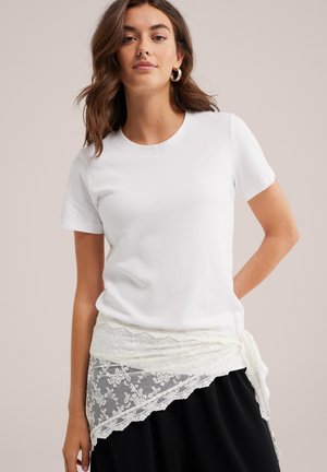 Femme aux cheveux bruns ondulés portant une chemise blanche à manches courtes, des boucles d'oreilles créoles dorées et une jupe noire avec une ceinture en dentelle blanche nouée à la taille.