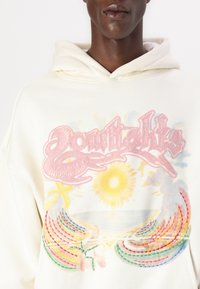 Felpa crema con testo ricamato rosa, grafica di un sole e motivi arcobaleno sulla parte frontale. Tessuto morbido e vestibilità rilassata.