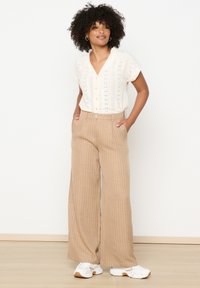 White knitted top with a V-neck and short sleeves, paired with beige striped wide-leg trousers and white sneakers. Wooden floor background.