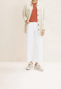 Hellsbeige Button-Up-Jacke über einem rot-weißen gestreiften Hemd, kombiniert mit weit geschnittenen, weißen, verkürzten Hosen und beigen Sneakers.