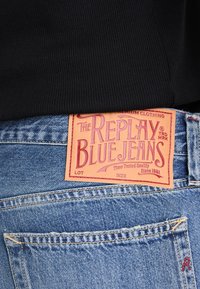 Vue de dos d'un jean bleu en denim avec une étiquette en cuir cousue indiquant « The Replay Blue Jeans » et un haut noir côtelé rentré à l'intérieur.
