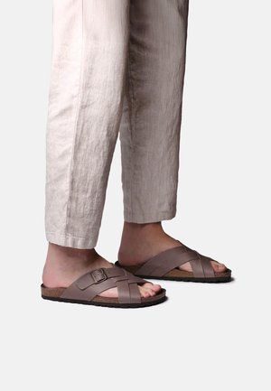 Jalad, mis kannavad beežikaid lühikesi pükse ja pruune nahast sandale, millel on ristuvad rihmad ja pandel, valgel taustal.