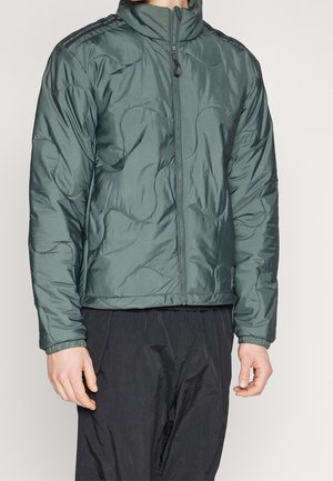 Homme portant une veste matelassée à fermeture éclair vert foncé avec des poignets élastiques et un pantalon large noir sur un fond uni.