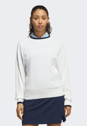 Jeune femme portant un pull blanc à manches longues avec des poignets côtelés et une jupe bleu marine fendue devant, debout devant un fond uni.
