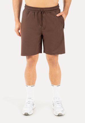 Mann trägt knielange braune Shorts mit elastischem Bund, weiße Sportsocken und weiße Sportschuhe, steht mit entspannten Händen.