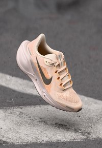 Helle rosa Nike Laufschuhe mit schwarzem Swoosh-Logo, in der Luft über Asphalt mit weißen Straßenmarkierungen.
