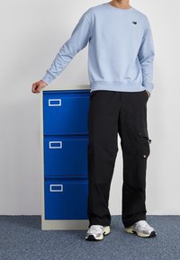 Světle modrá mikina s žebrovanými manžetami, černé cargo kalhoty s kapsami a šedé tenisky, stojící vedle modrého kancelářského kontejneru.