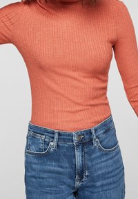 Rippentop mit langen Ärmeln in Koralle, mit hohem Kragen, kombiniert mit hoch geschnittenen blauen Jeans aus Denim mit Knopfverschluss und Vordertaschen.