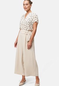 Roomkleurige blouse met een V-hals, gecombineerd met ruime beige broeken met een riemdetail. Lichte schoenen maken de outfit compleet.