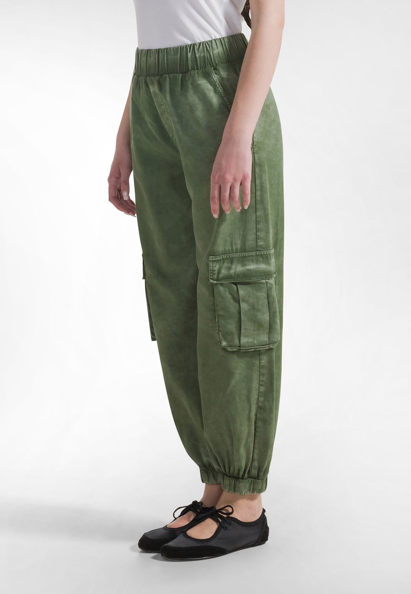 Spodnie cargo w jasnym oliwkowym kolorze z elastycznym pasem, elastycznymi mankietami i dwoma bocznymi kieszeniami na przechowywanie. Dopełnieniem stylizacji są czarne płaskie buty.